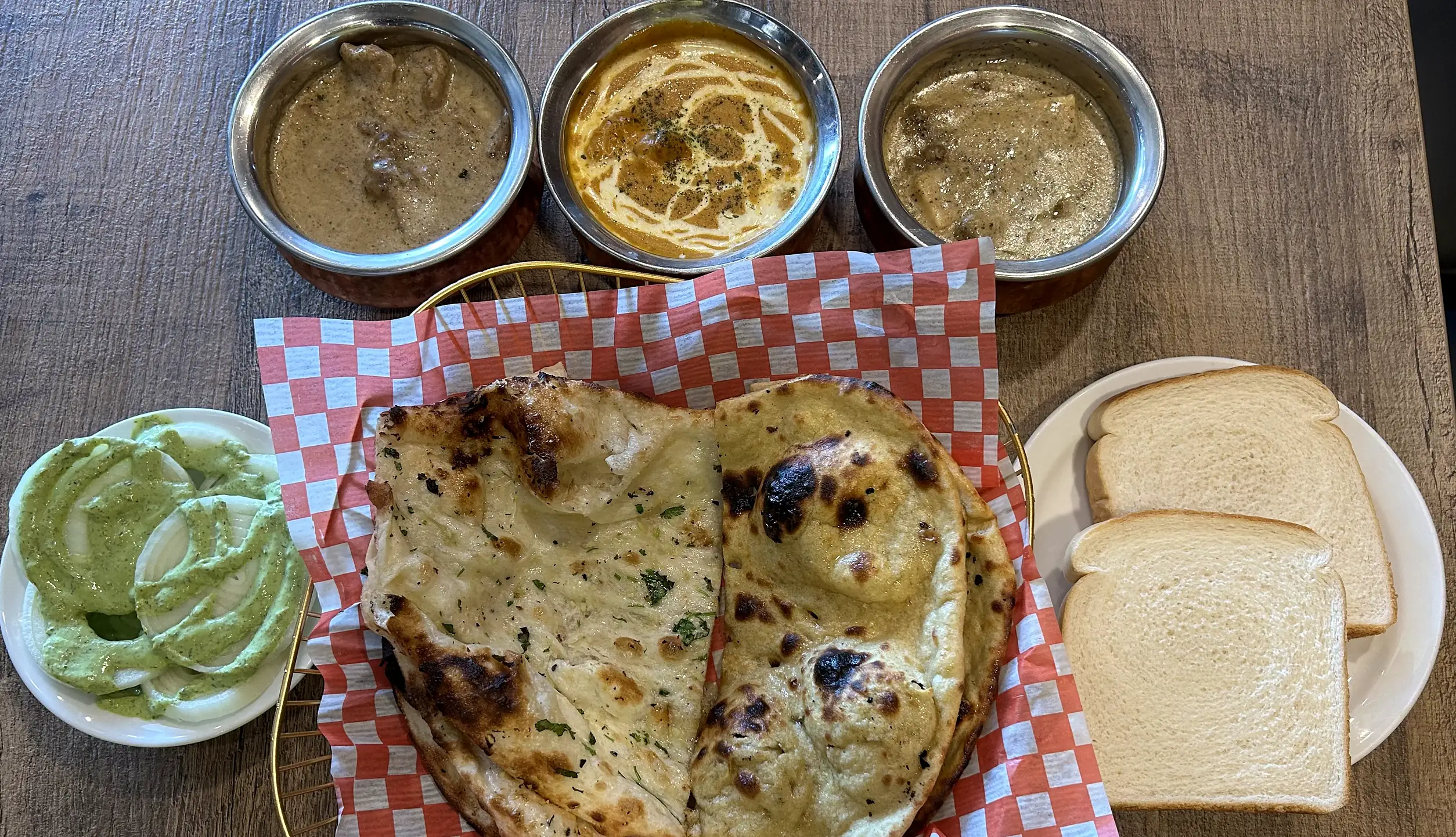 Three curries in steel bowls with butter naan in a checkered paper-lined basket, white bread slices and mint chutney on a wooden table