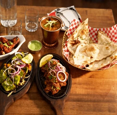 Two tikka sizzlers with onion rings and lemon beside glazed chicken, a copper cup of curry and a naan basket