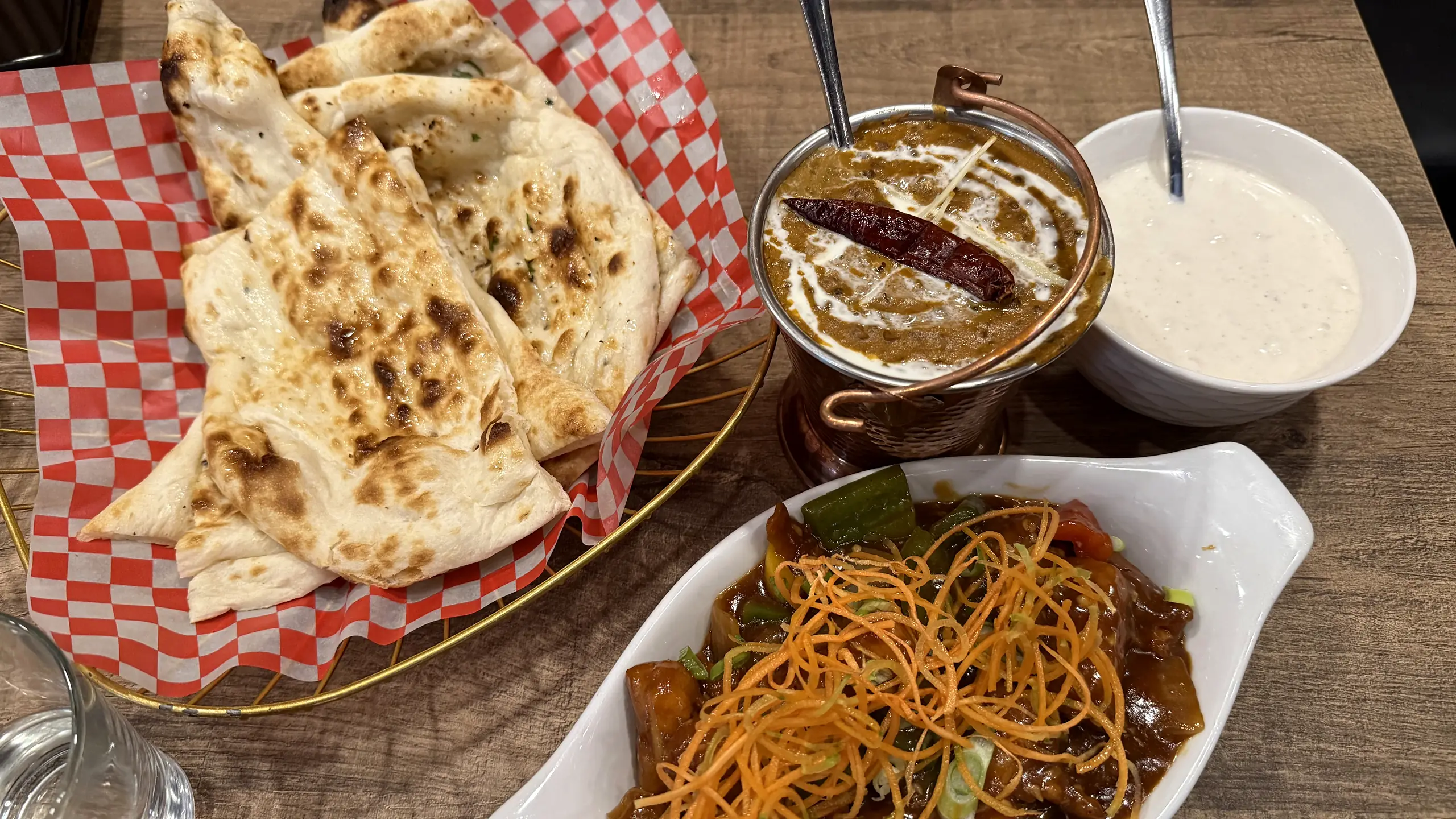 Butter naan in a checkered paper-lined basket with dal makhani in a copper cup, raita and a saucy vegetable dish topped with shredded carrot
