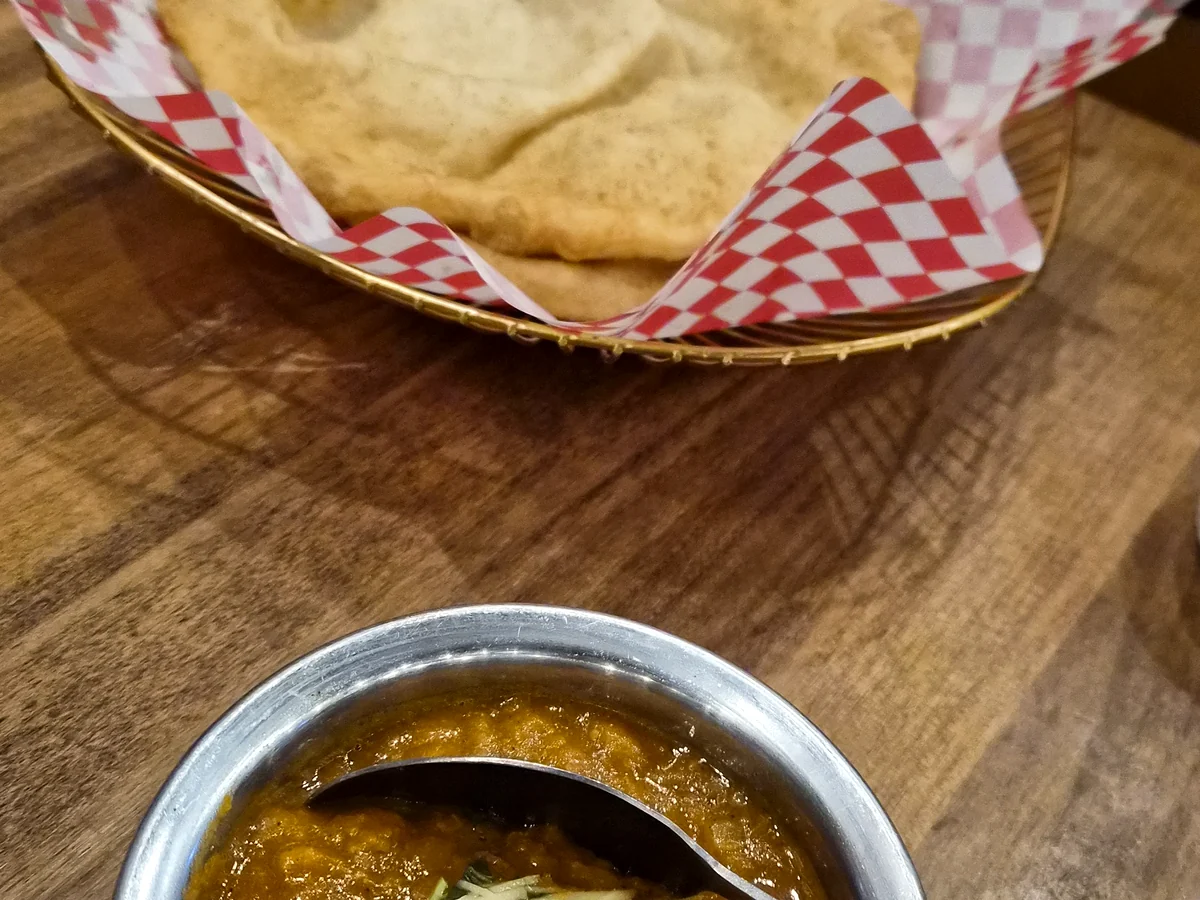 Chole garnished with ginger and coriander in a steel bowl, with bhature in a checkered paper-lined basket behind