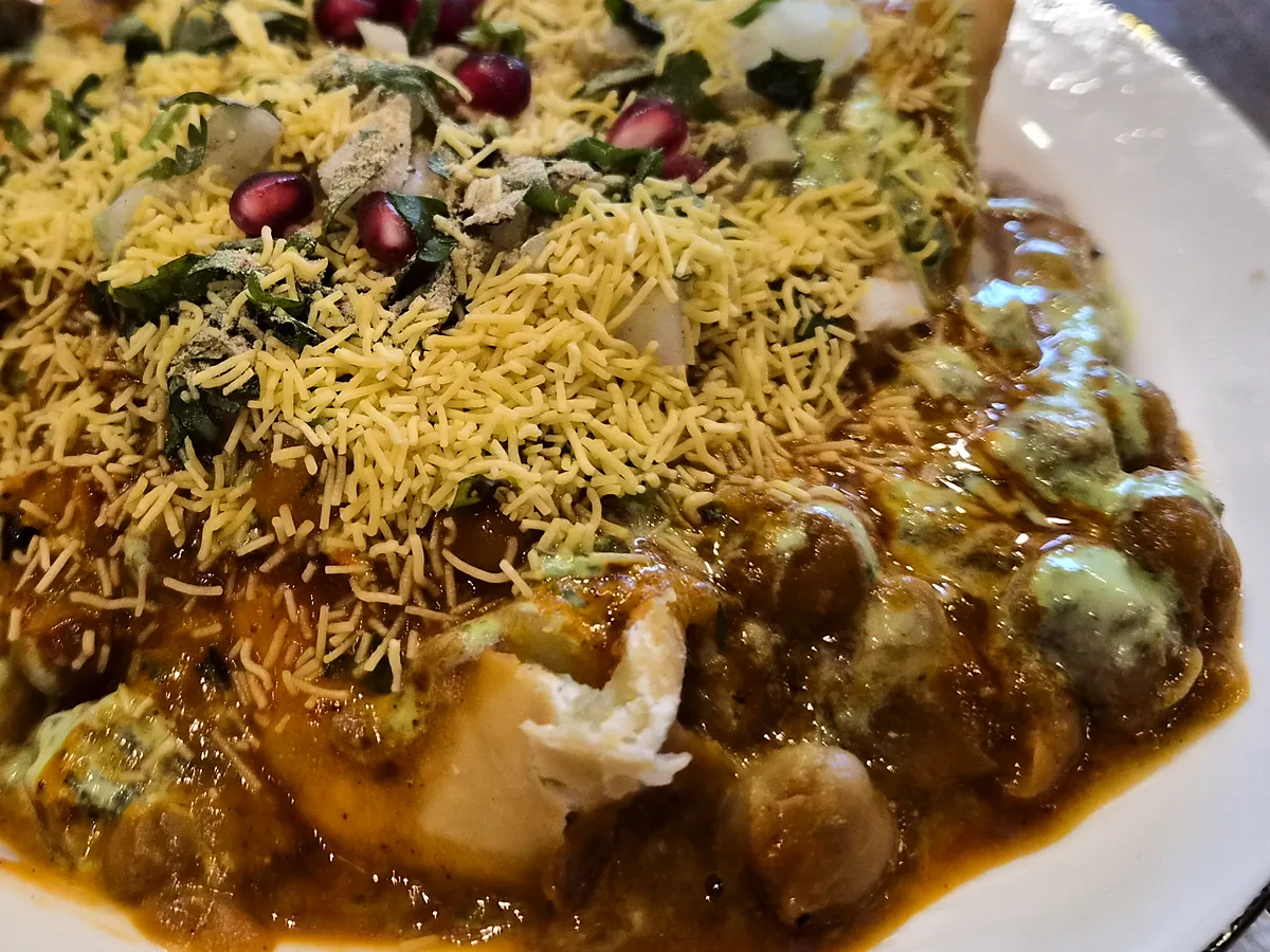 Samosa chaat with a broken samosa under spiced chickpeas, mint chutney, sev and pomegranate seeds on a white plate