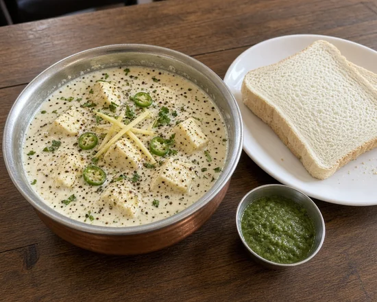 Cream paneer in a black-pepper cream gravy with green chilli rings in a steel bowl beside white bread